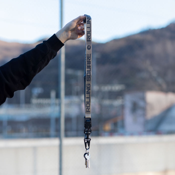 Rolling Square Lanyard - Digital ID & QR Lost Mode - Silver Fog - Charcoal black Rolling Square lanyard with repeating logo text and metal clip held against blurred outdoor background. - Rolling Square